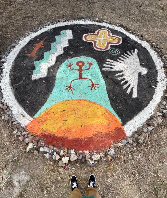 A man stands before a sand painting created by Indigenous artists on the ground. The painting is circular and framed by rocks. A bird, cross, sun, and more colorful symbols are depicted.