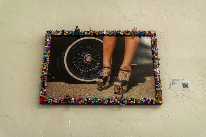 A photo is framed on a wall, and the frame is decorated with miscellaneous items like gems. Pictured is the feet of a woman, tucked into clear high heels; one foot has an "S" tattoo, and the other has a "D" tattoo.