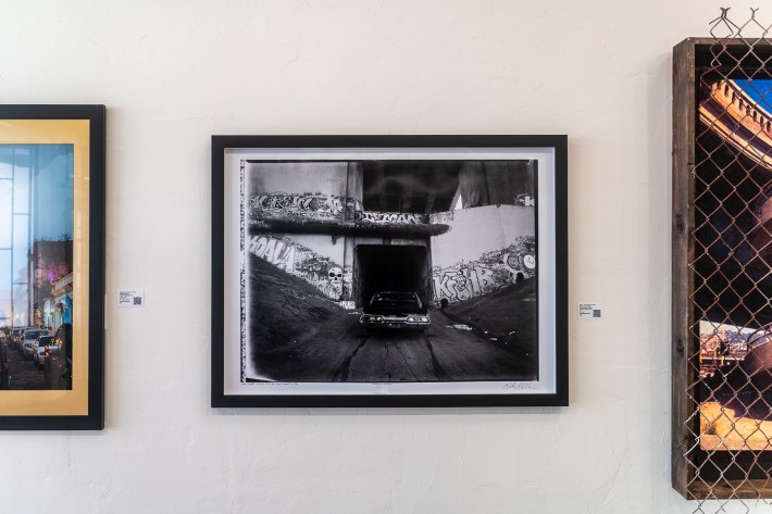 Photos are hung on a wall's art gallery. The center photo depicts an iconic L.A. tunnel in black and white. A black 1967 Chevy Impala car is driving into the tunnel.