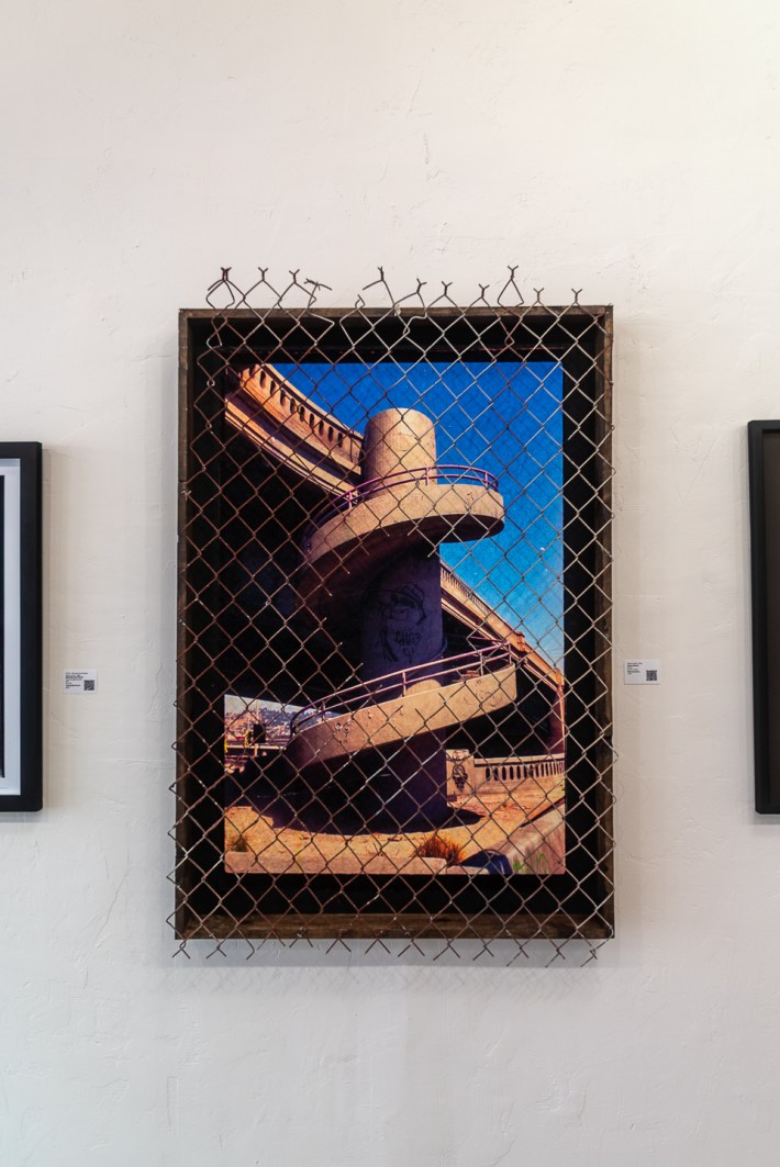 An image depicting an industrial, outdoor spiral staircase decorated with graffiti is framed behind a metal fence.