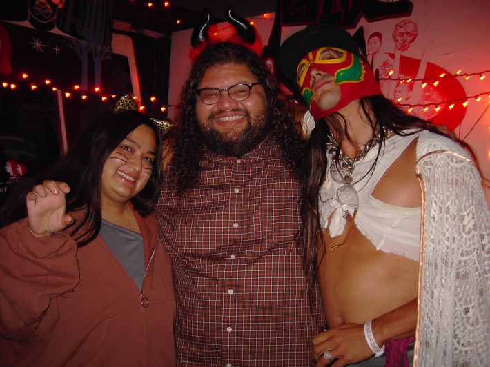 A woman dressed as a cheetah cat, a man dressed as a devil, and man dress as Lucha Libre pose together.