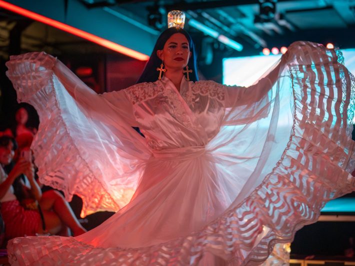 A woman in a white gown raises its hems to show the detailed design. She is balancing a candle on her head, and she is onstage.