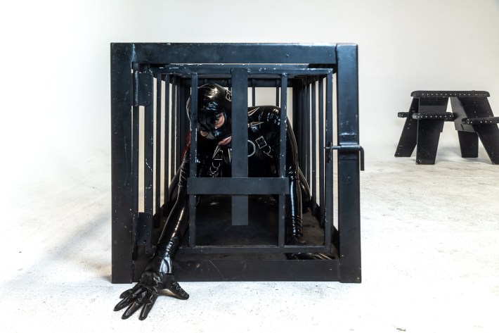 A woman in a full-body latex suit poses from within a BDSM cage. A standing horse furniture piece is in the background.