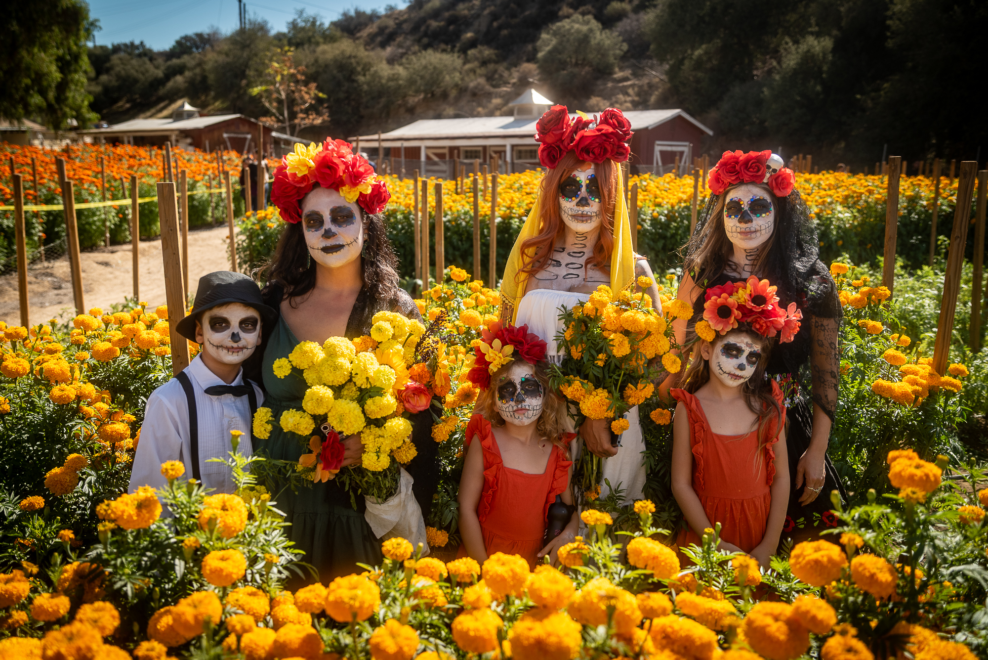 three women and three children pose in front of flowers while wearing outfits celebrating Dia de los Muertos