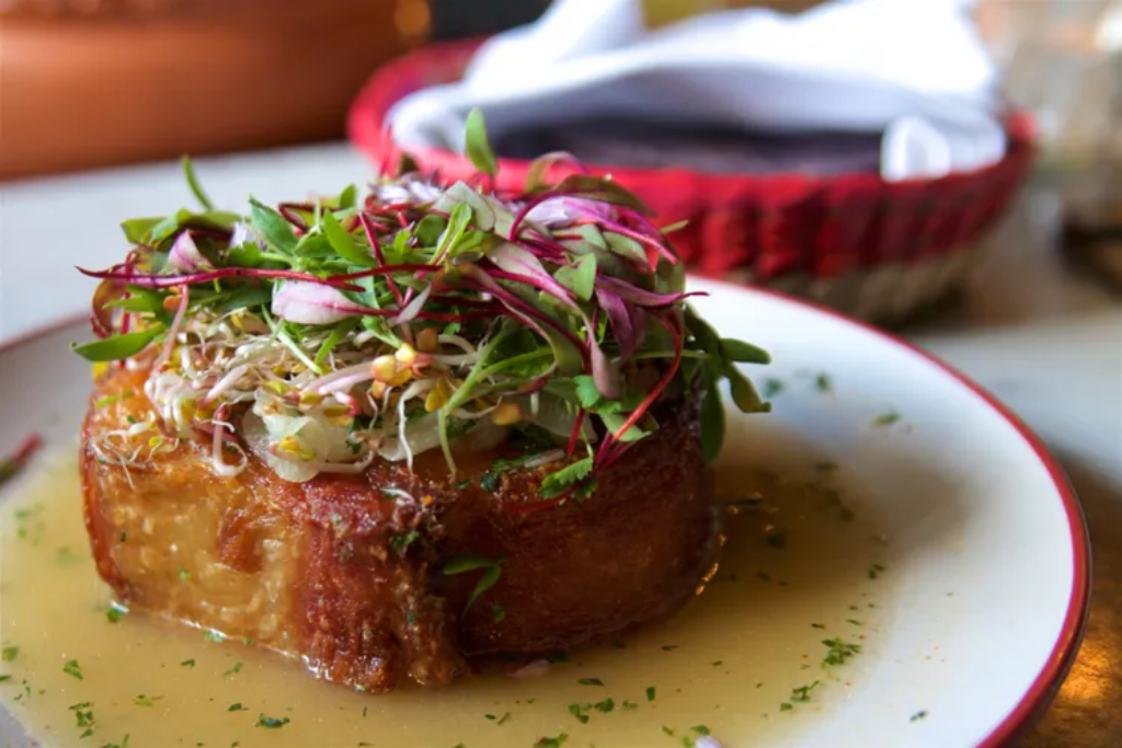 A round slab of chicharron pork topped with microgreens in a pool of green liquid on a white plate, with a tortilla basket behind it