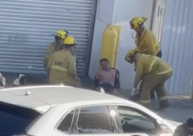 A man slouches against a wall after being shot. Firefighters are around him.