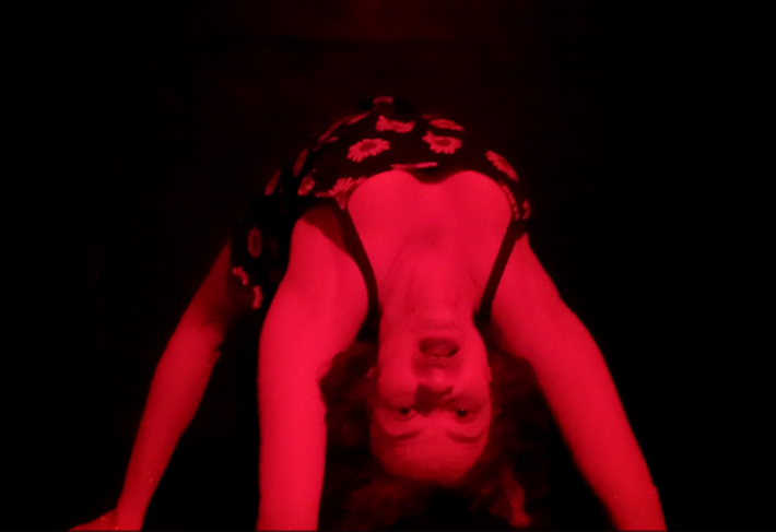 A woman hangs upside down on all fours, in red light, looking menacingly at the camera