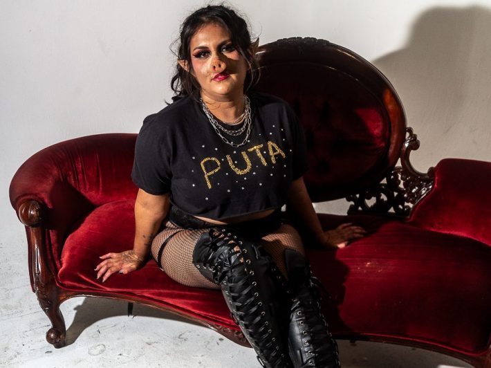 A woman wearing a prosthetic pig nose, a black tee shirt reading "PUTA," and thigh-length black boots poses on a red chaise in front of a white background.
