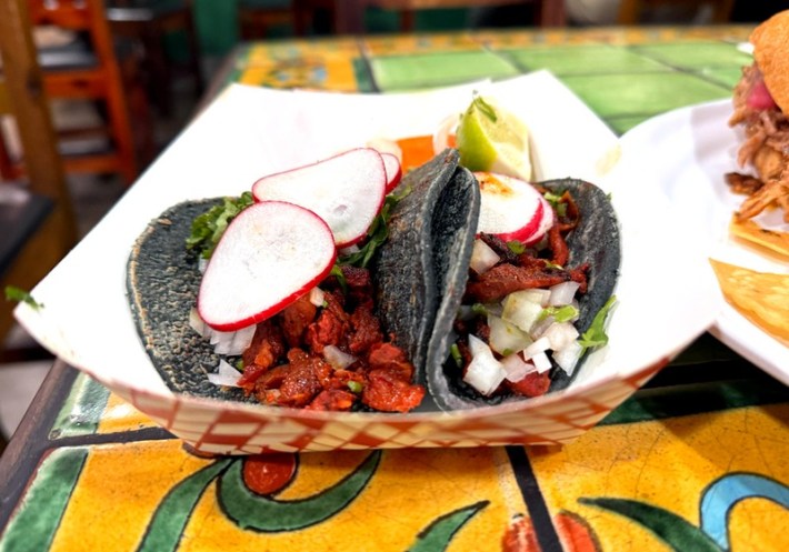 a taco with beef shoulder and longaniza on blue corn tortillas topped with radishes in a paper holder
