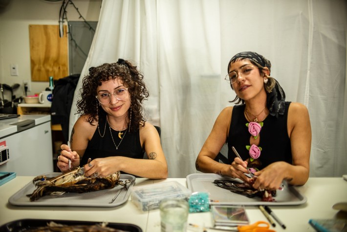 Two women smile at the camera despite their hands still working on their taxidermy projects.