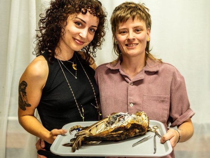 Two smiling women pose closely together, both holding onto a tray with a dead bird ready to begin the taxidermy process.