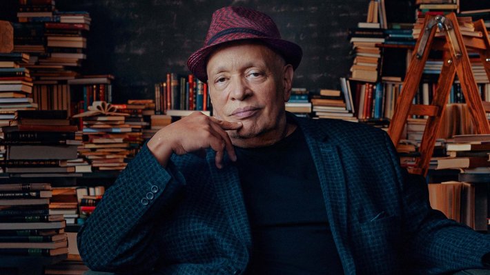 A man in a fedora sits in front of a stack of books.