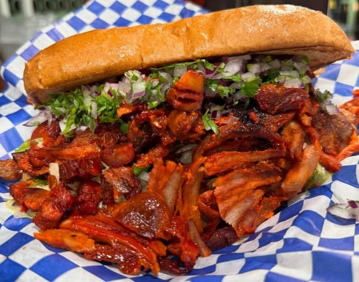 A torta with al pastor spilling out of the front onto a blue-and-white checkered paper