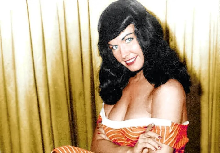 A woman with dark hair smiles into the camera. She is wearing a low-cut, orange dress with white abstract stripes.