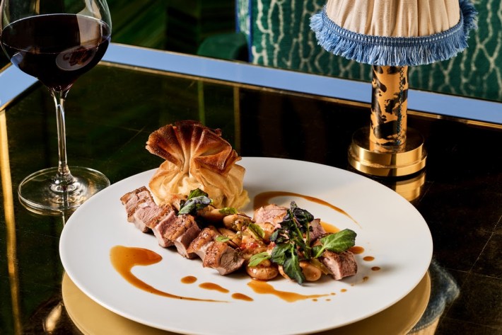 A dish of sliced, seared Rohan duck breast next to a potato dumpling and greens, on a white plate, between a lamp and glass of red wine