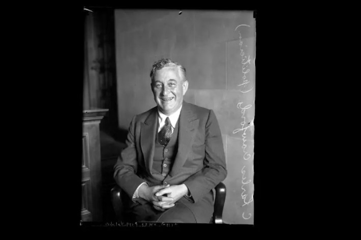 An older man sits in a suit, smiling into the camera.
