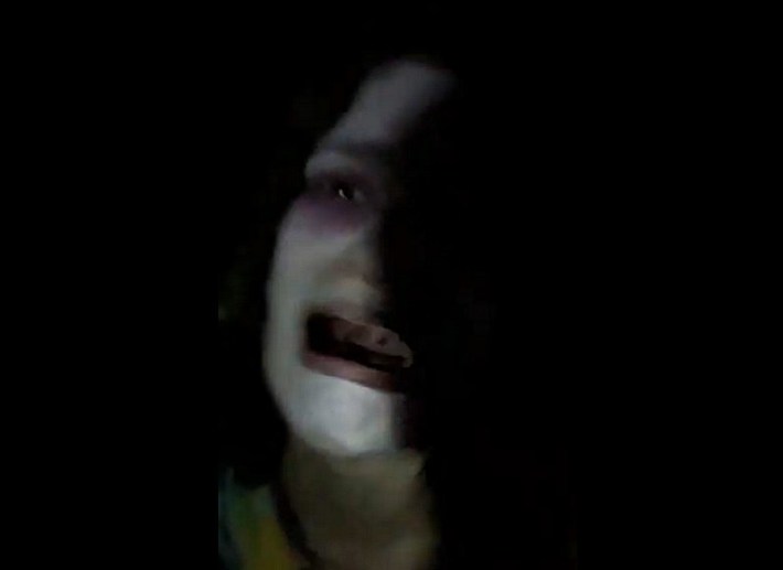 A woman in white zombie face paint cries at the camera, half obscured by darkness