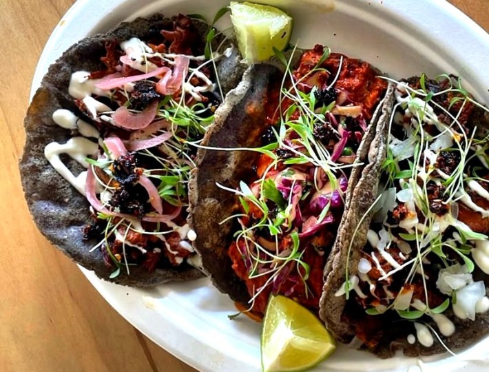 Three tacos with crema on bluie corn tortillas with micro greens on top, on a plate next to 2 wedges of lime