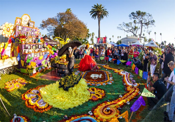 A crowd celebrates Dia de los Muertos, watching a performer in celebratory makeup and attire.