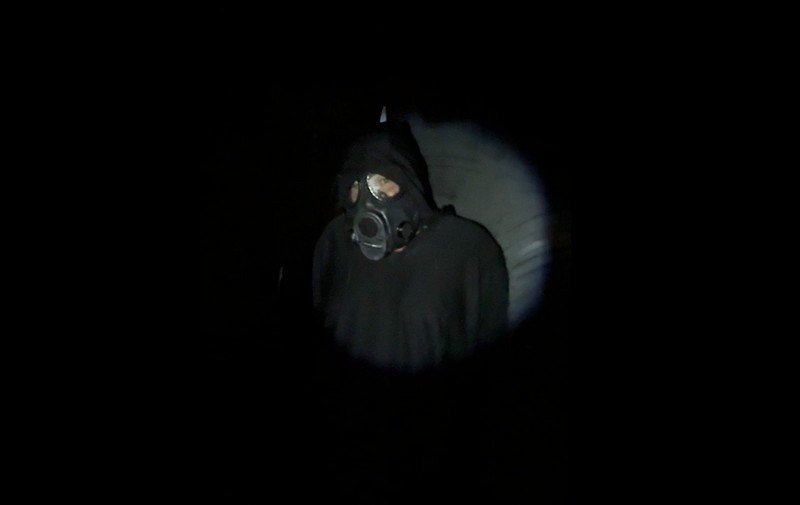 A hooded man in a gas mask in a flahslihgt's spotlight in a dark space