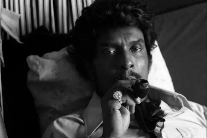 A man with curly hair is resting on a pillow with a pipe in his hand and held to his lips.