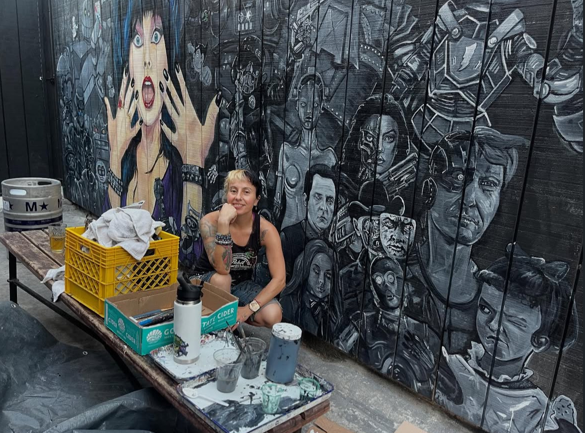 A woman poses in front of a mural of faces she painted.