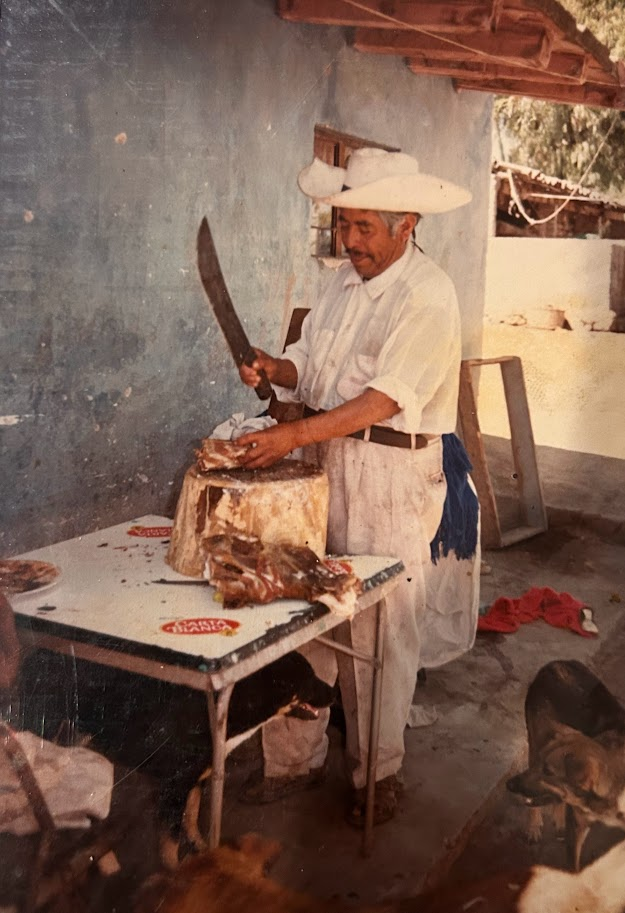 A man in a cowboy hat cuts a slab of meat on a tree trunk slab.