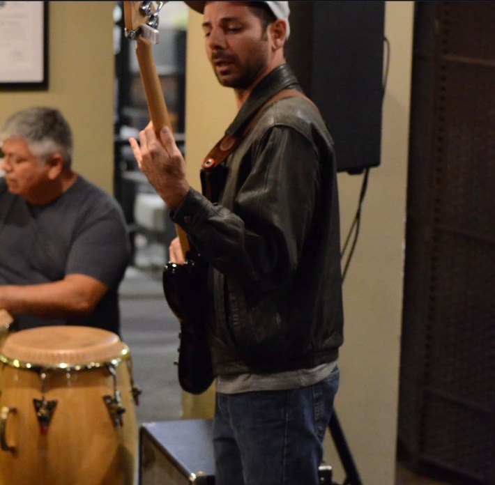 A man in a cap and jacket plays a bass guitar.