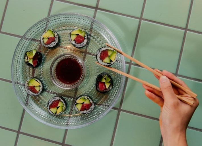 A glass plate of beef kimpab with someone's hand holding one roll in chopsticks, soy sauce in the middle