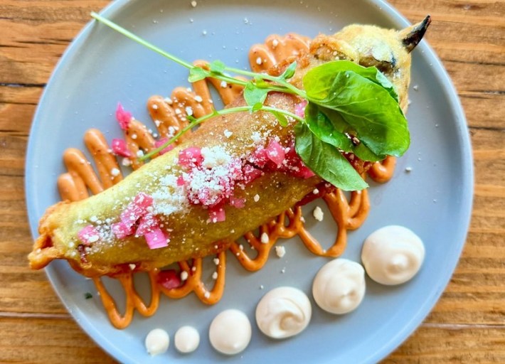 Hatch chile relleno on a light blu plate surrounded by dots of white sauce and squiggles of an orange sauce