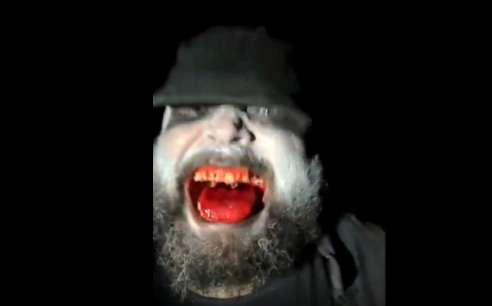 A bearded man with a white painted face, dark eyes, and a bloody mouth and teeth lunges at the camera