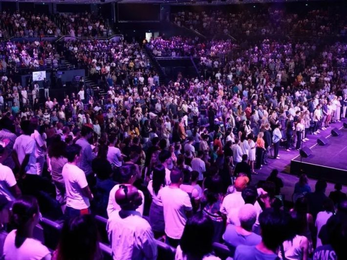 A crowd of people in a stadium stand beneath purple lights.