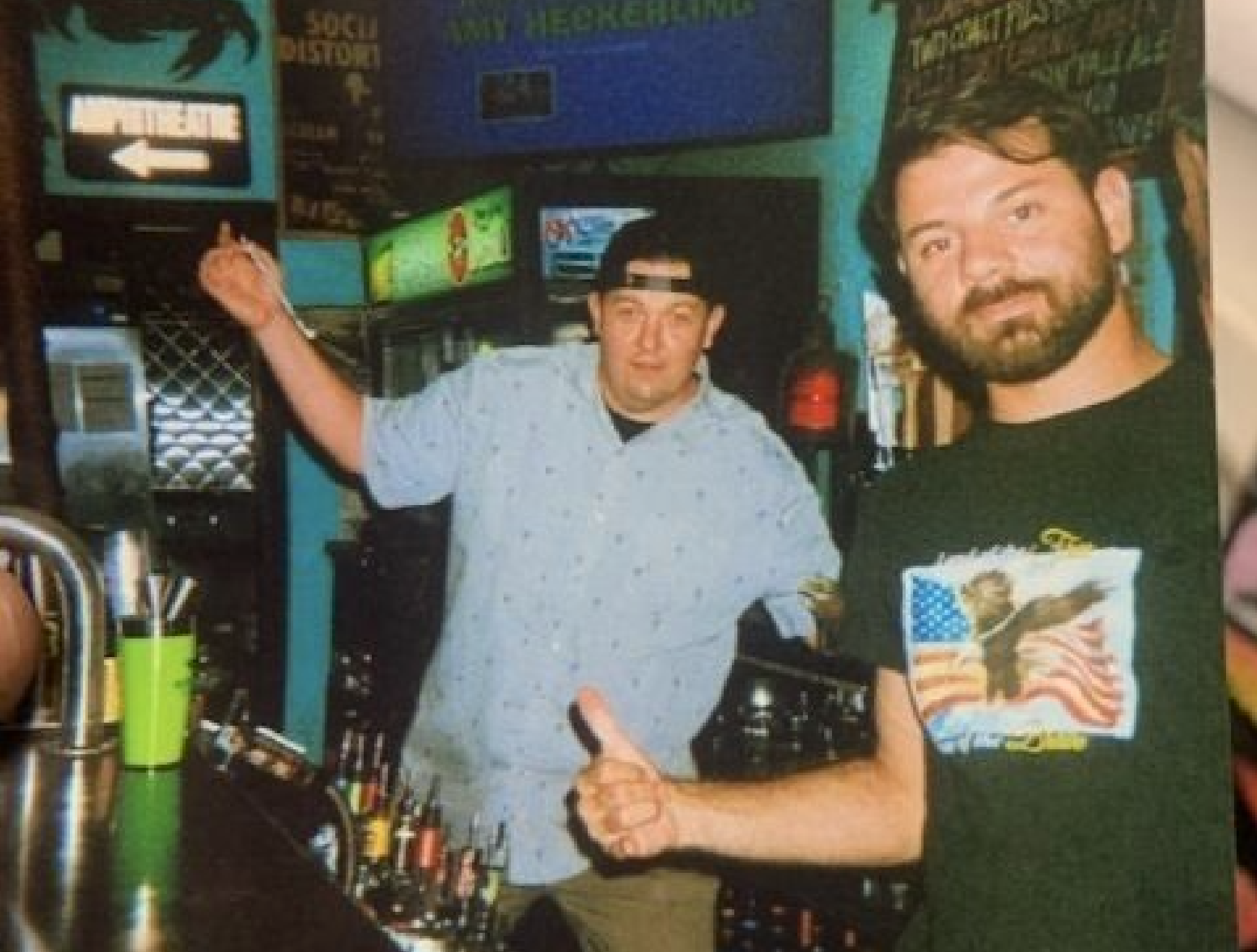 Two men pose at a bar, one giving a thumbs up.