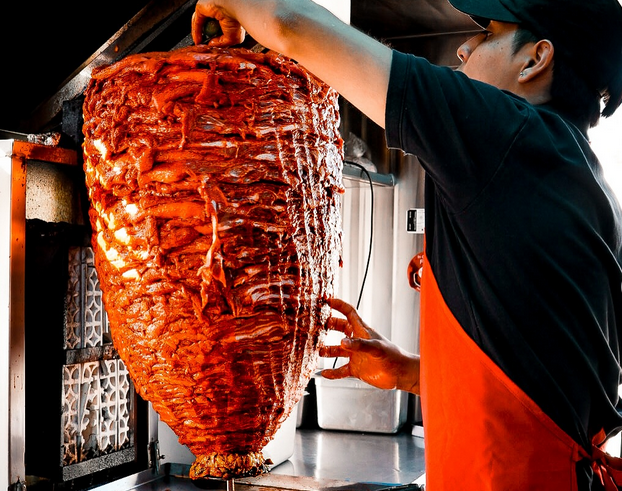A man in a red apron secures a big trompo of al pastor before its heat source