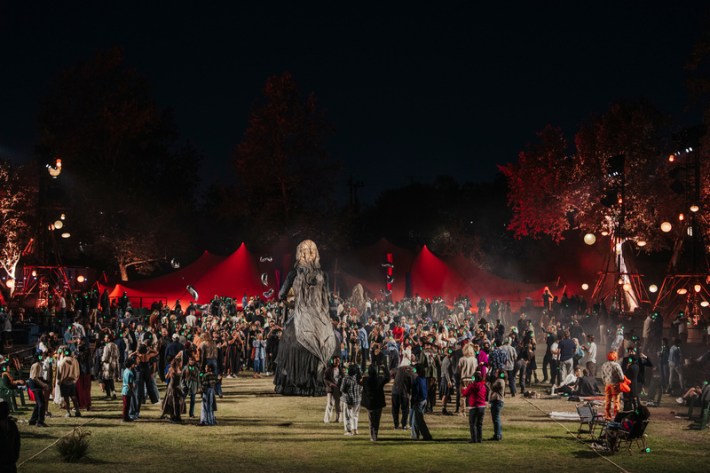 A crowd dances around a giant puppet in the open air with red lights behind it