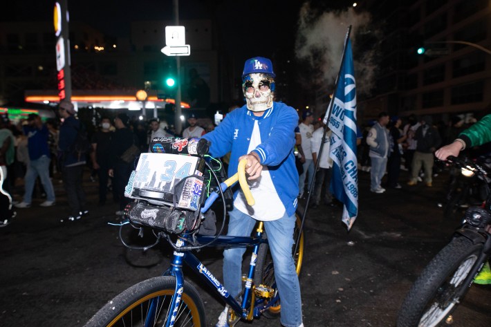 a man in a skull mask stands posed with his bicycle