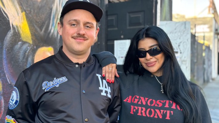 a man with a mustache and a woman with sunglasses smile next to each other