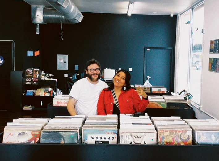 a man in a white shirt and a woman in a red jacket smile and pose together from within rows of vinyl records