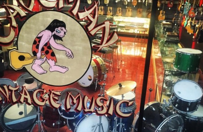 a logo on a glass wall shows a caveman with bangs holding a guitar. beyond the wall, there are various drum kits, guitars, and more instruments on display
