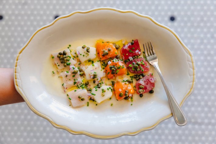 A plate of crudo featuring four different cuts of colorful fish, topped with capers, next to a fork on a white plate
