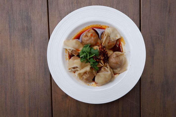 A white plate full of Chinese dumplings in a red chile oil sauce and topped with cilantro