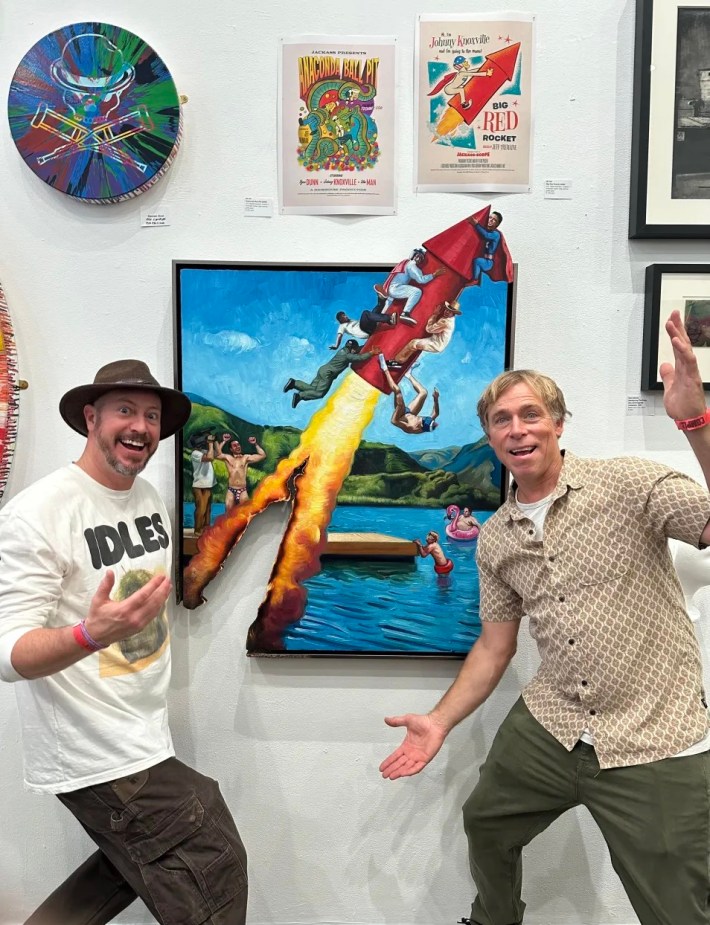 two men pose in front of an art piece depicting a rocket of TNT