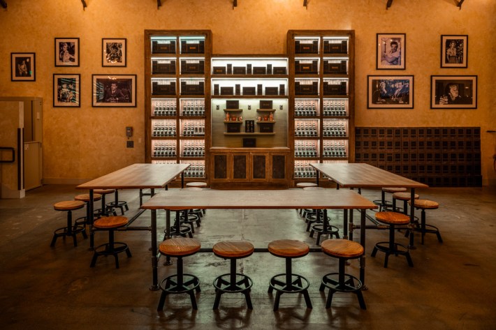 A tasting room, at a distillery, with a u=shaped network tables and chairs, and shelves stocked with bottles and boxes, framed photos bookending them