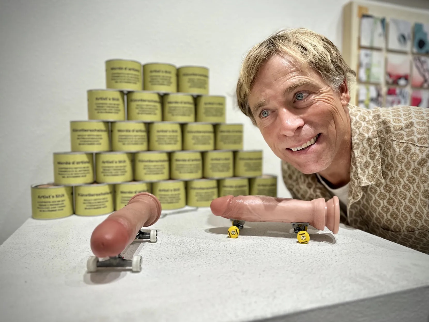 a man smiles next to a sculpture of skateboards where the boards have been replaced with realistic dildos resembling penises