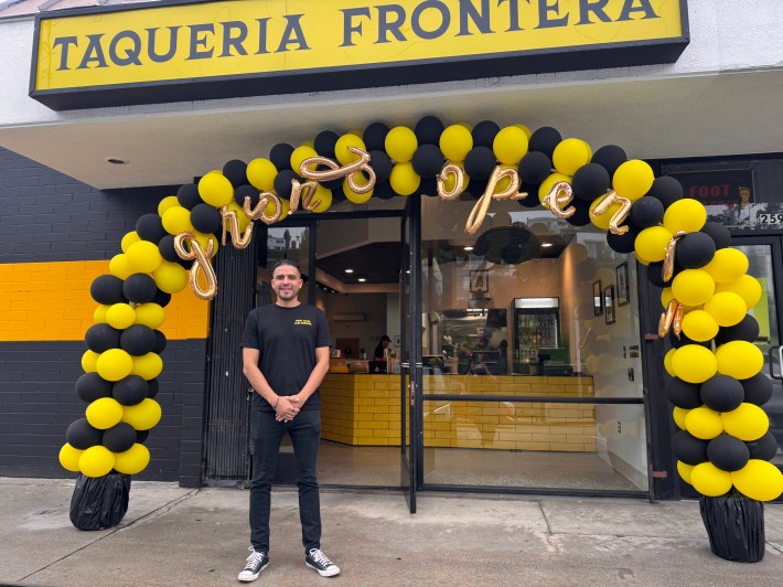 a man stands in front of a storefront. a balloon arch reads "grand opening"