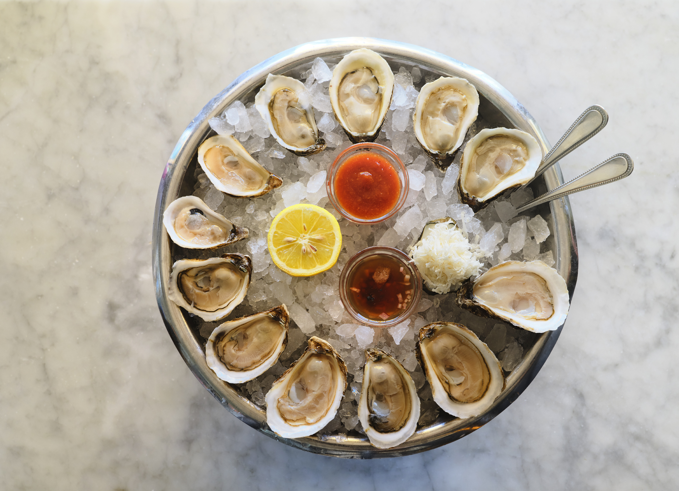 A platter of twelve oysters arranged in a circle on a bed of ice, with lemon and sauces