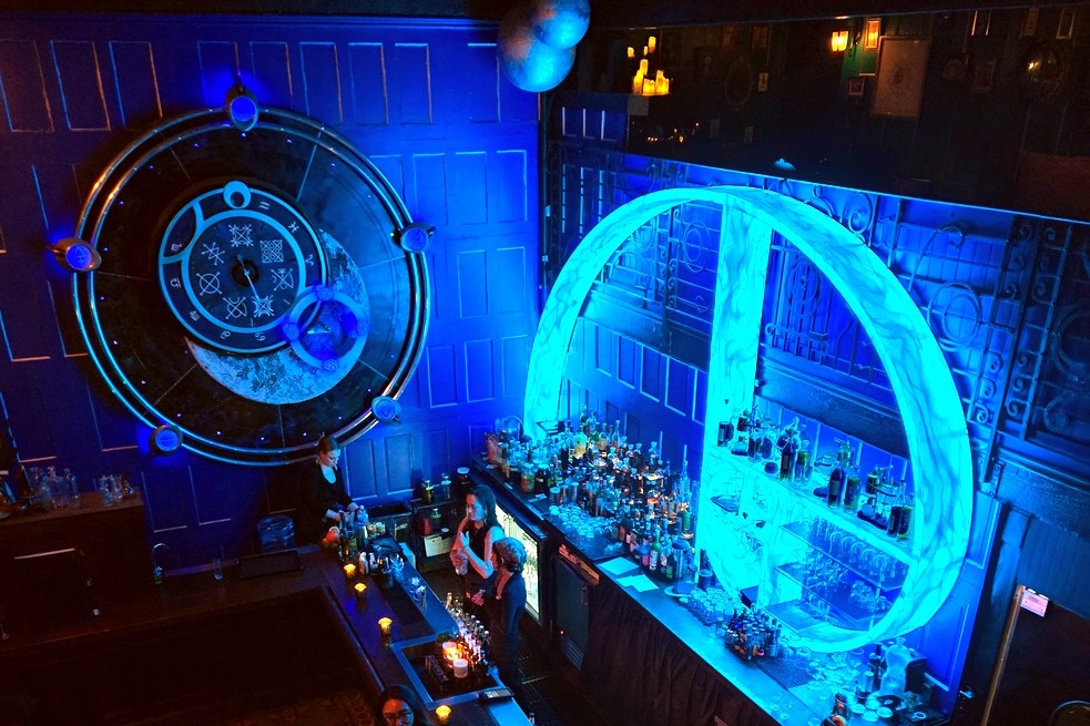 A bar seen from above with a huge snail-shaped blue neon artwork behind it, and bartenders serving customers