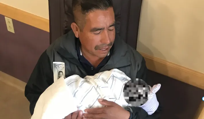 A seated man with a mustache holds a swaddled infant, looking at her