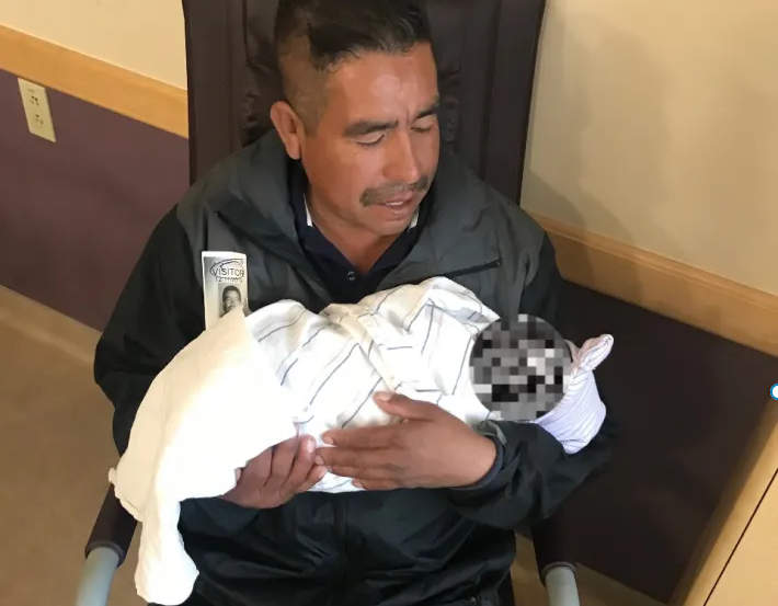 A seated man with a mustache holds a swaddled infant, looking at her