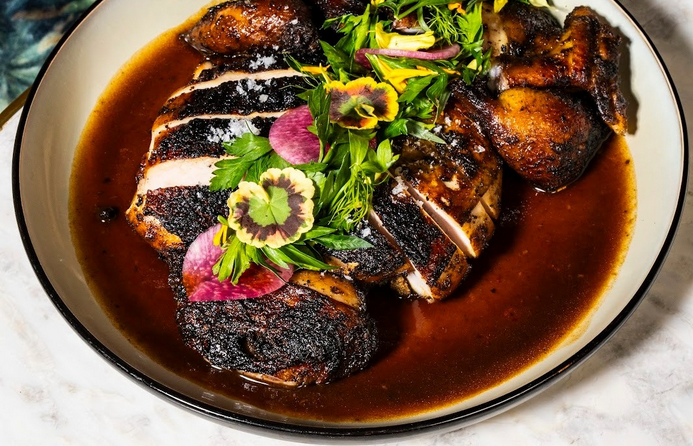 A plate of sliced, pimento-smoked jerk chicken, topped with herbs and sitting in a dark red jus.